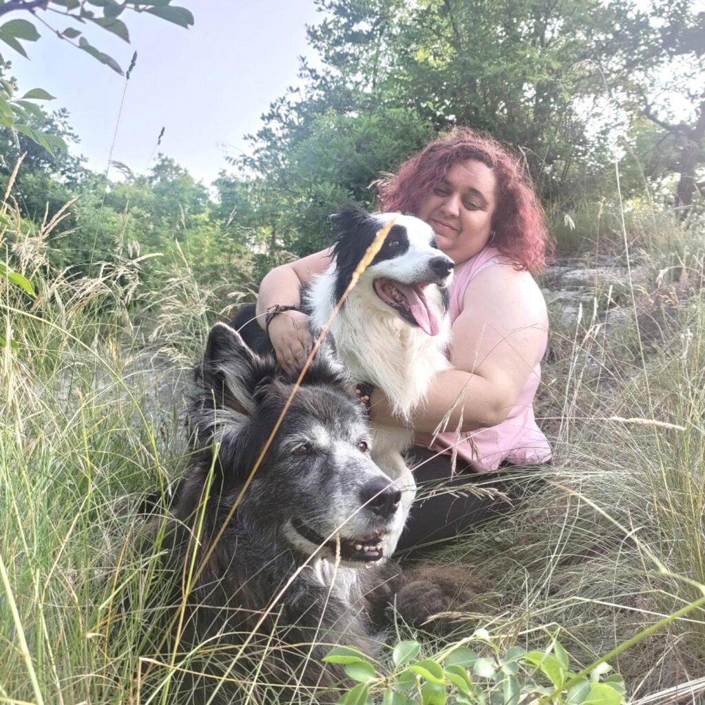 L'istruttrice cinofila Gloria Levrini seduta nell'erba alta insieme ai suoi cani Tesla e Neko, in un momento di serena condivisione all'aperto, in un attimo del percorso relazionale e professionale.