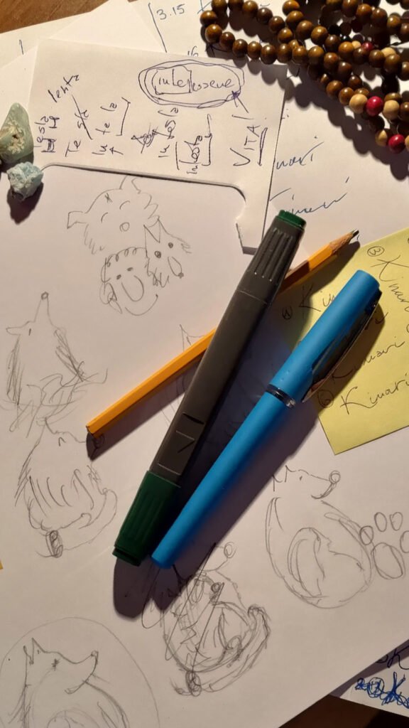 Vista dall'alto di fogli di carta con schizzi a matita e prove di calligrafia per il logo Kinari - Intessere vita. Sopra i fogli sono appoggiati una penna azzurra, un pennarello grigio, una matita, alcune pietre grezze di larimar e una mala di legno di sandalo.