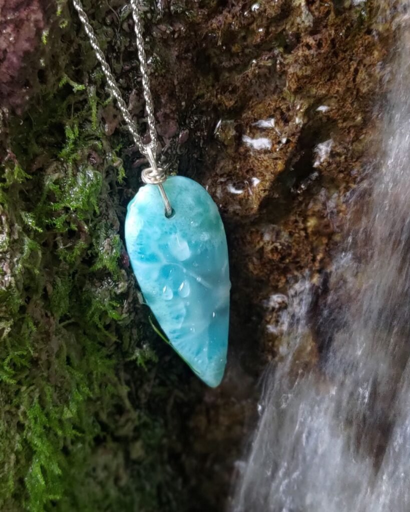 Un ciondolo di pietra Larimar azzurra con venature bianche è appeso a una catenina d'argento, adagiato su una roccia umida con muschio verde accanto allo scorrere di una cascata d'acqua.